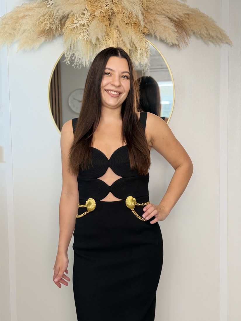 A model wearing a black occasion dress with gold detailing, photographed in the salon.